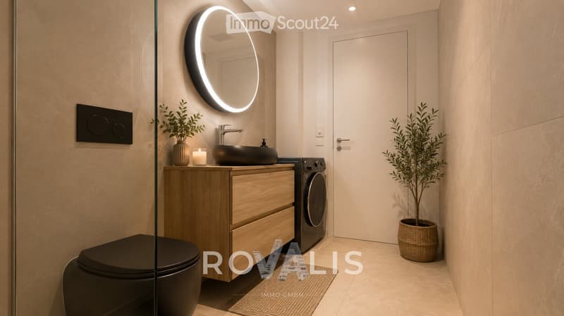 Modern bathroom with a sleek black sink, round illuminated mirror, wooden vanity, toilet, and washing machine. Features include a potted plant, candle, and recessed lighting.
