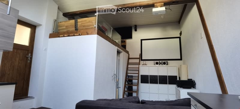 Modern living room with a dark grey sofa, a black table, a white storage unit, a large screen mounted on the wall, speakers, and a wooden ladder leading to an upper level.