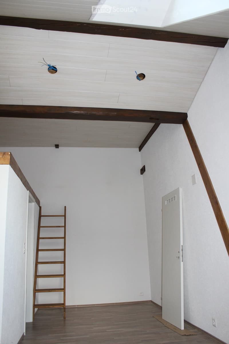 Empty attic room with wooden floor, white walls, exposed wooden beams, and a small ladder leading to the ceiling.
