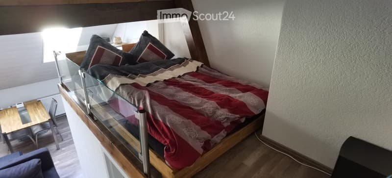 A cozy bedroom with a wooden bed, red and white patterned bedding, a small wooden table, and a chair near a large window with a view.