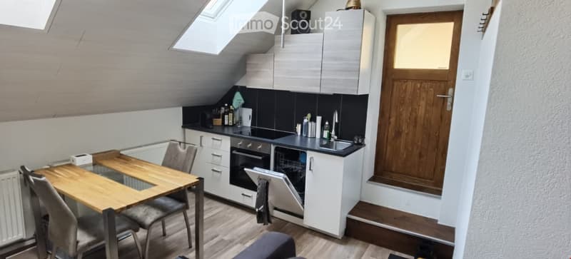 Modern kitchen with white cabinets, black countertops, a wooden dining table with chairs, and built-in appliances including an oven and dishwasher. The space features a skylight and a wooden door.