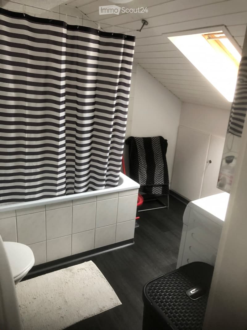 Modern bathroom with dark wooden flooring, white tiled bathtub, black and white striped shower curtain, toilet, washing machine, and a small stool.