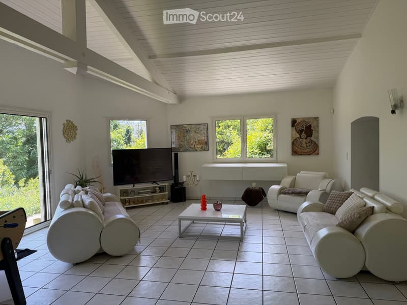 Bright living room with white walls, tiled floor, and large windows. Features a white sectional sofa, coffee table, TV, side table, and decorative art pieces.