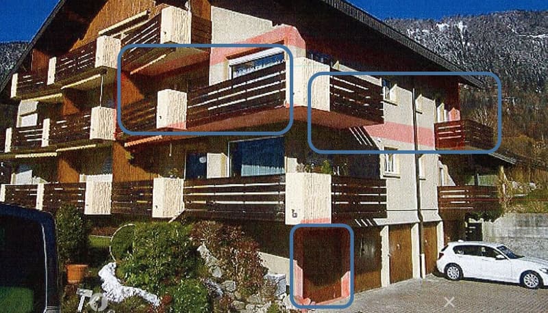 A multi-story residential building with balconies, wooden facades, and a parking area. The structure features a mix of modern and traditional architectural elements, with visible balconies and a white car parked in the foreground.