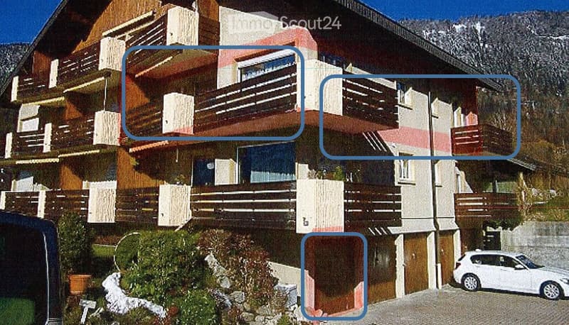 A multi-story residential building with balconies, wooden facades, and a parking area. The structure features a mix of modern and traditional architectural elements, with visible balconies and a white car parked in the foreground.