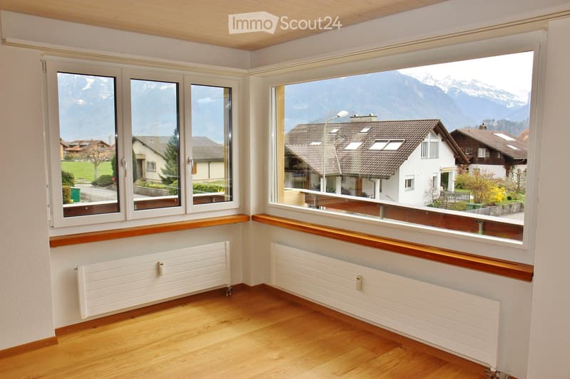 Empty room with wooden flooring, white walls, large windows offering a scenic mountain view, and radiators beneath the windows.