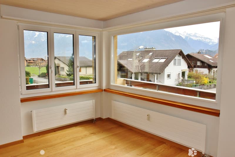 Empty room with wooden flooring, white walls, large windows offering a scenic mountain view, and radiators beneath the windows.