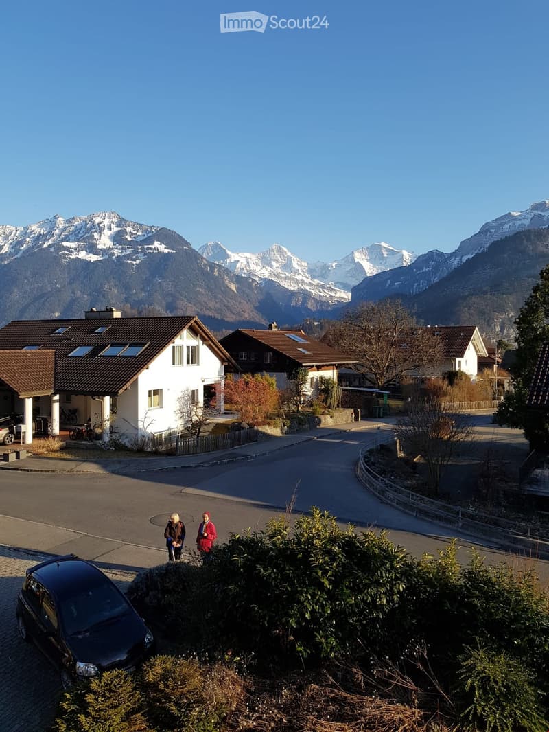 Residential street with houses, parked cars, and snow-capped mountains in the background.