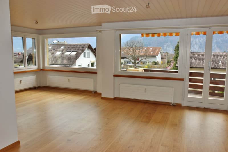 Empty room with wooden flooring, large windows, radiators, and a view of the surrounding area.
