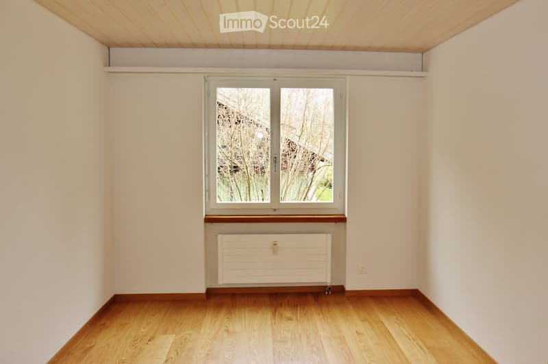 Empty room with light wooden flooring, white walls, and a large window with a radiator below it.