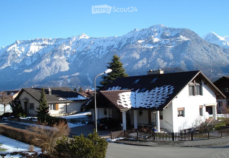 Two-story house with white exterior, dark roof, and snow-covered mountains in the background.