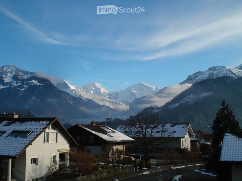 A row of houses with snow-covered roofs and surrounding snow-capped mountains in the background.