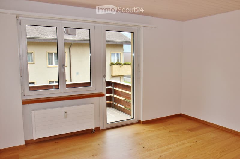 Empty room with wooden flooring, white walls, large windows, and a sliding glass door leading to a balcony.