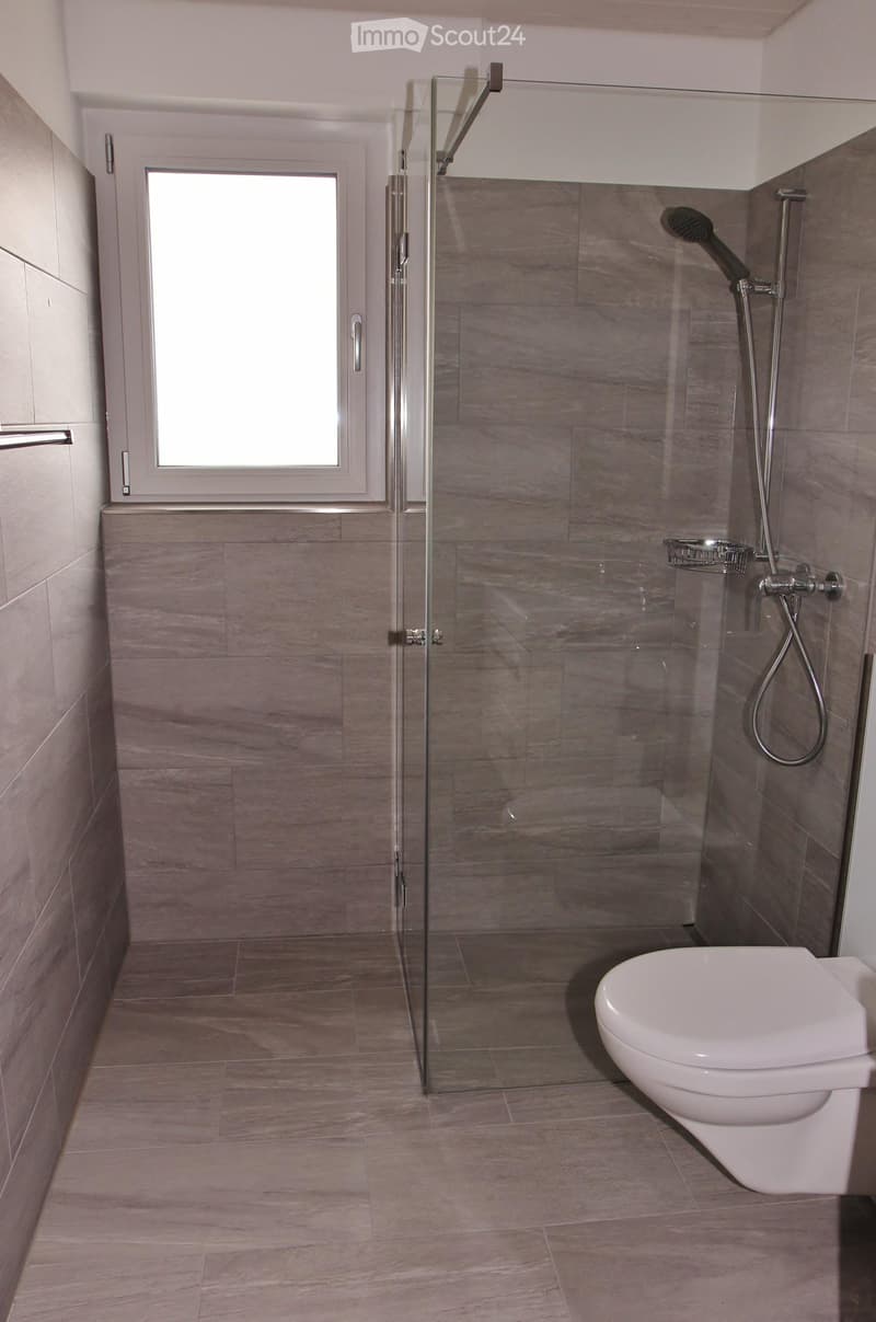 Modern bathroom with a glass-enclosed shower, a toilet, and a window providing natural light. The walls and floor are tiled in a light brown color.