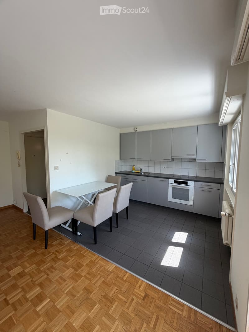 Modern kitchen with grey cabinets, white dining table, four chairs, and built-in oven. The floor is a mix of dark tiles and light wooden parquet.
