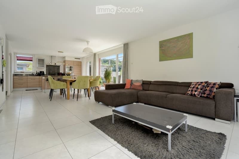 Modern living and dining area with a brown sectional sofa, coffee table, dining table with chairs, kitchen area with cabinets, and a large window with a view of greenery.