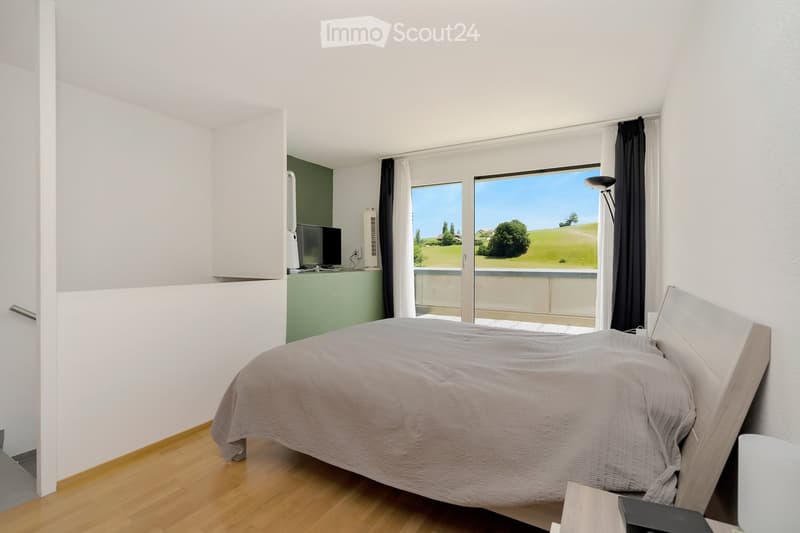Modern bedroom with a large bed, nightstand, lamp, and a balcony overlooking a green landscape.