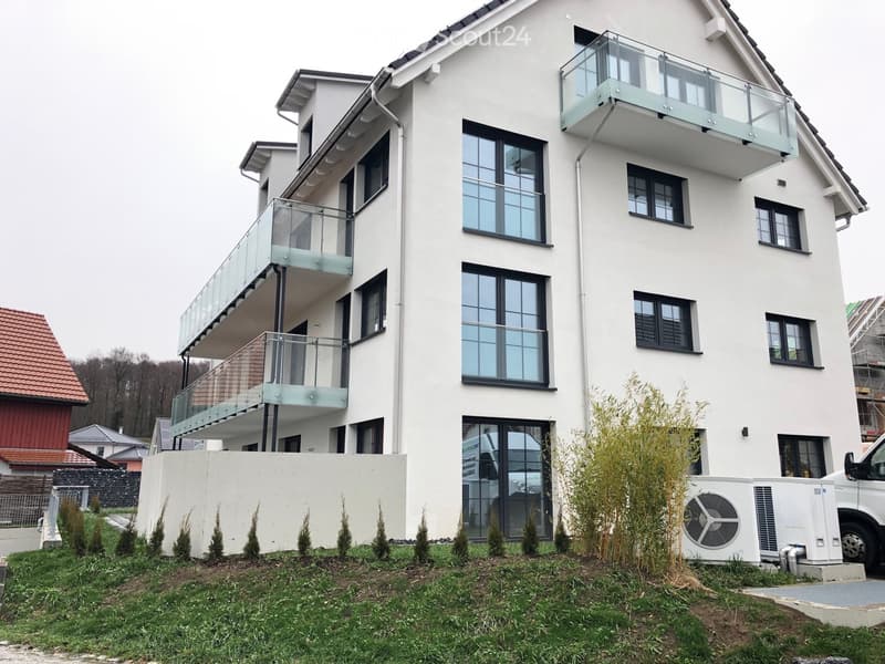 Modern two-story house with white exterior, large windows, and glass balconies. Features a well-maintained lawn and small shrubs in the front yard.