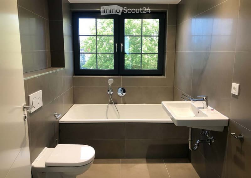 Modern bathroom with a bathtub, sink, toilet, and a large window offering a view of greenery outside.