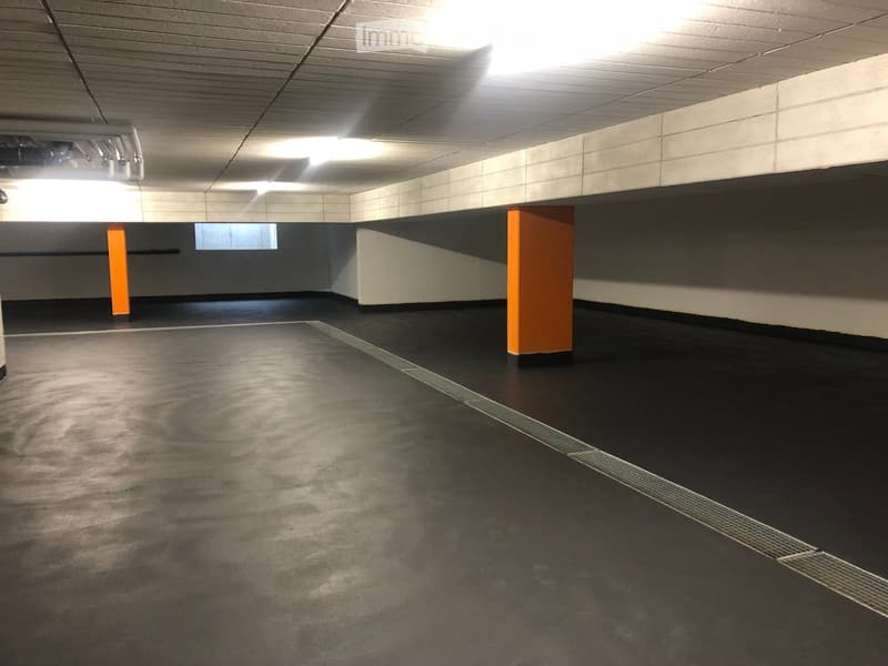 Underground parking area with black flooring, white tiled walls, and orange pillars. The space is empty with overhead lighting and a small window at the far end.