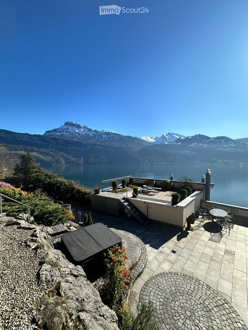 Outdoor terrace with a panoramic mountain and lake view, featuring a table, chairs, sun loungers, potted plants, and a hot tub.