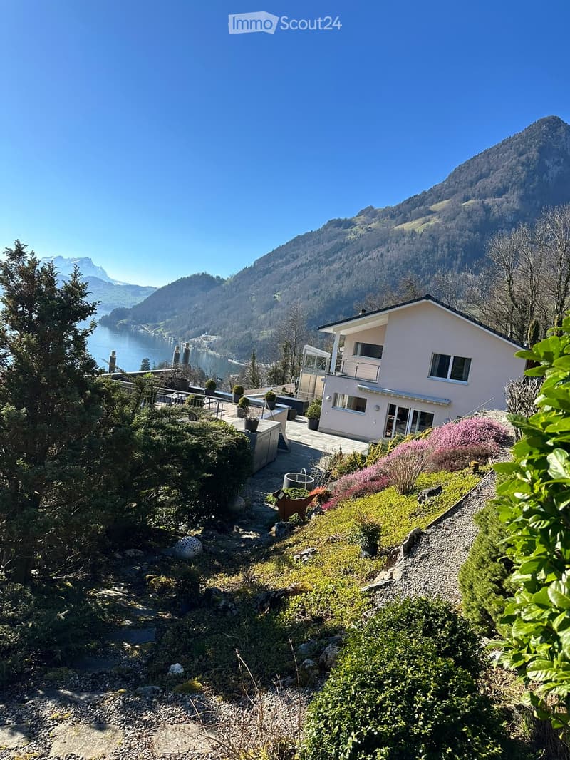 A modern house with a balcony overlooking a scenic lake and mountain landscape. The garden features various plants and a pathway leading to the house.