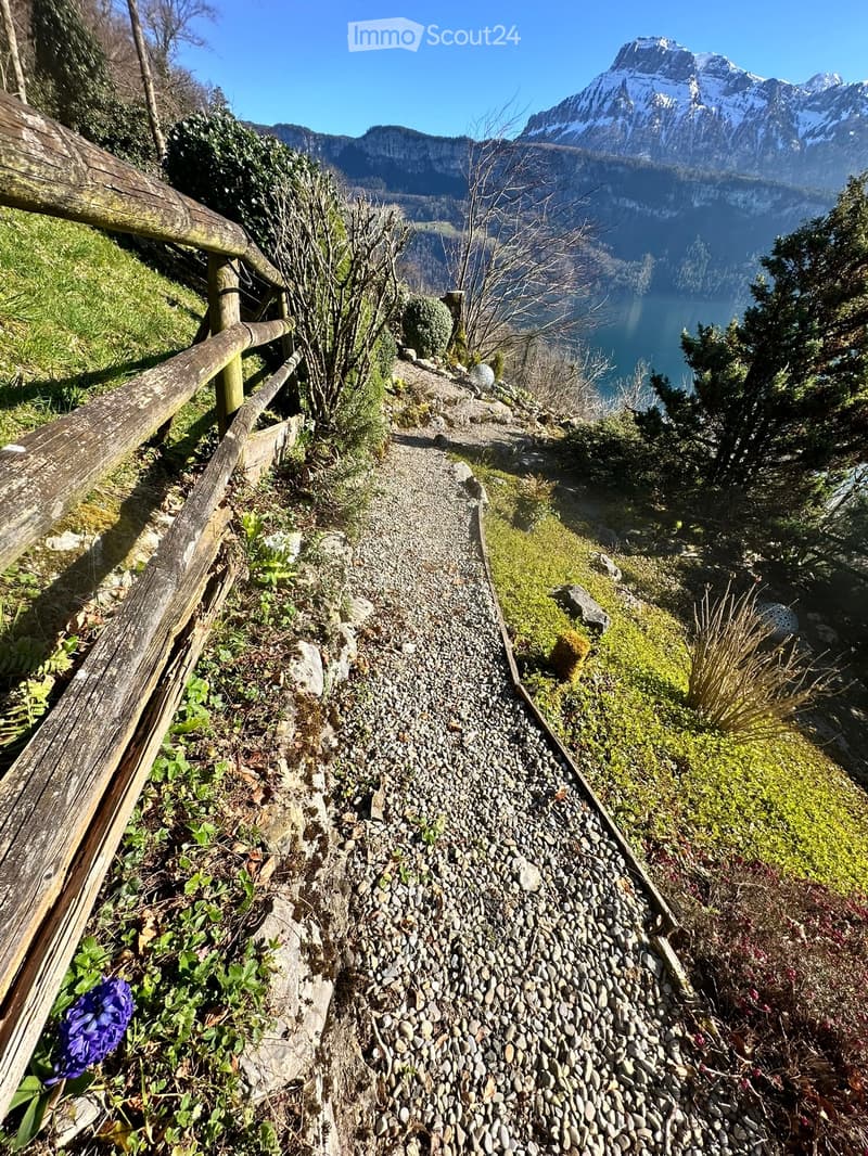 A narrow, gravel-covered pathway bordered by a wooden railing, leading up a hillside with sparse vegetation and a scenic mountain and lake view in the background.