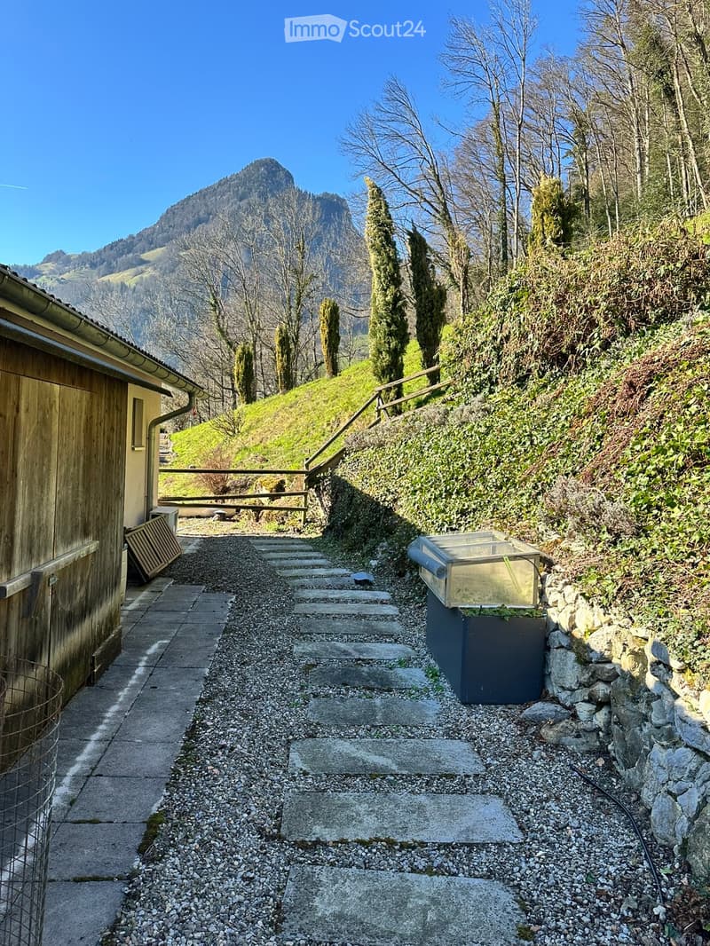 A stone pathway leads up a hillside, flanked by a wooden building on one side and a stone wall on the other, with a view of mountains in the background.