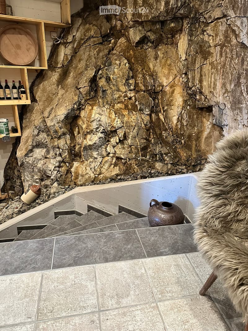 A room with a large stone wall, shelves holding bottles, a fur rug, and a decorative ceramic pot on the floor.