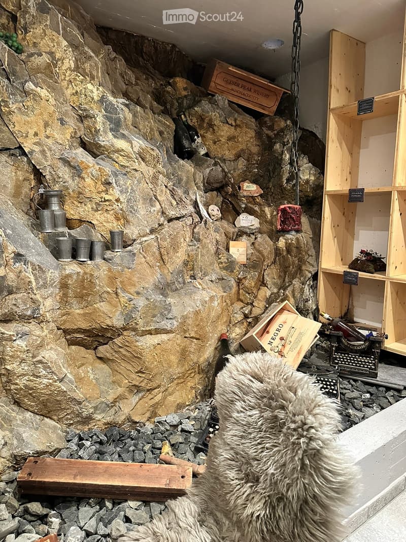 A cozy, rustic room with a stone wall backdrop, wooden shelves, wine bottles, wooden crates, a typewriter, and metal cups.