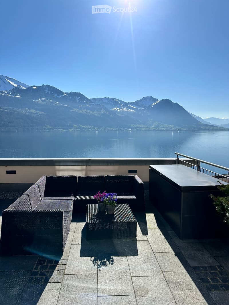 Balcony with a sofa, table, and flower pot, overlooking a lake and mountains.