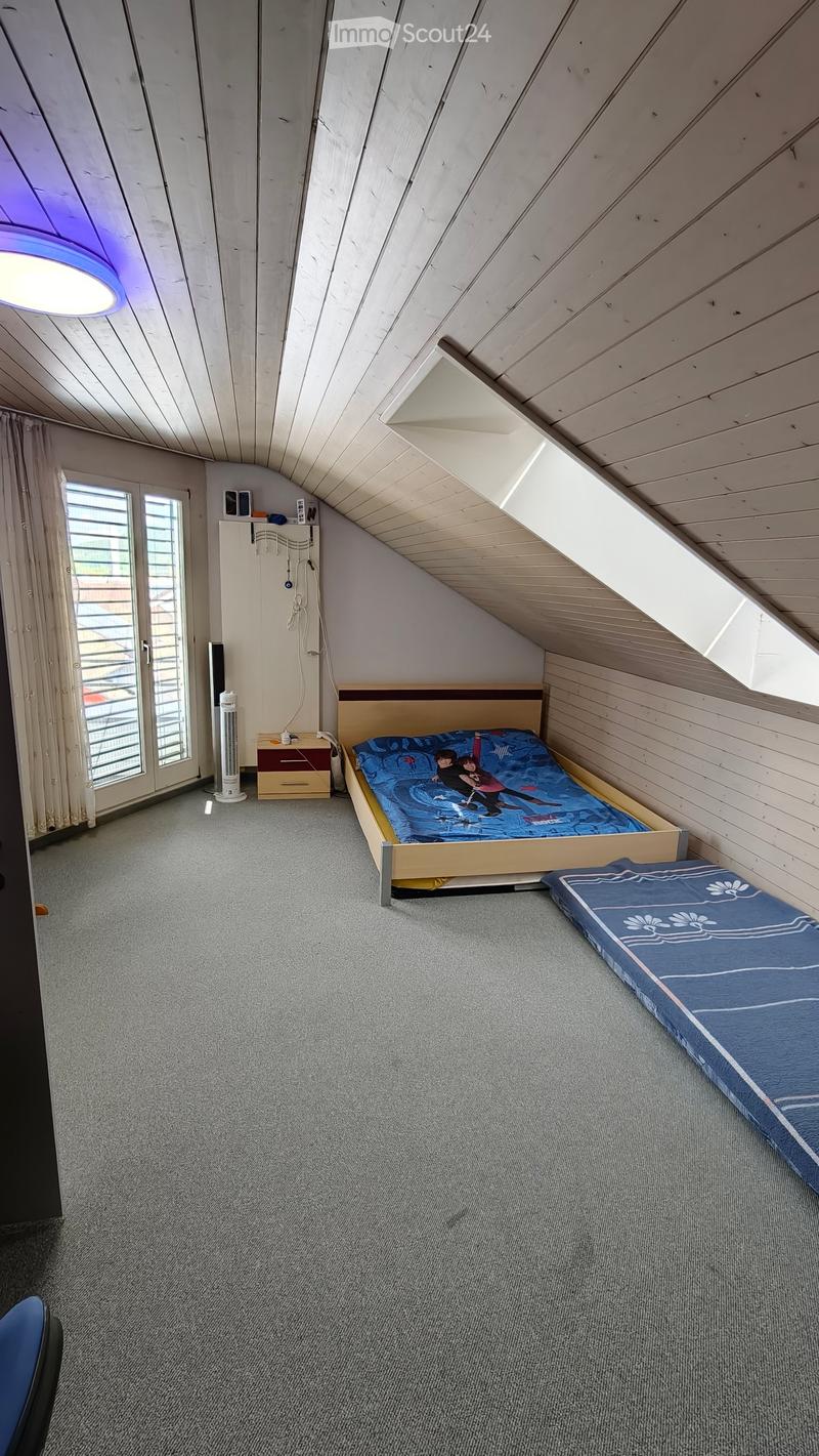 Cozy attic bedroom with slanted wooden ceilings, carpeted floor, and large windows. Features a single bed, nightstand, fan, and cabinet.