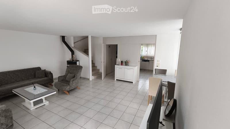 Bright living room with tiled floor, grey sofa, armchair, coffee table, dining area with chairs and table, television, and a small kitchen area visible in the background.