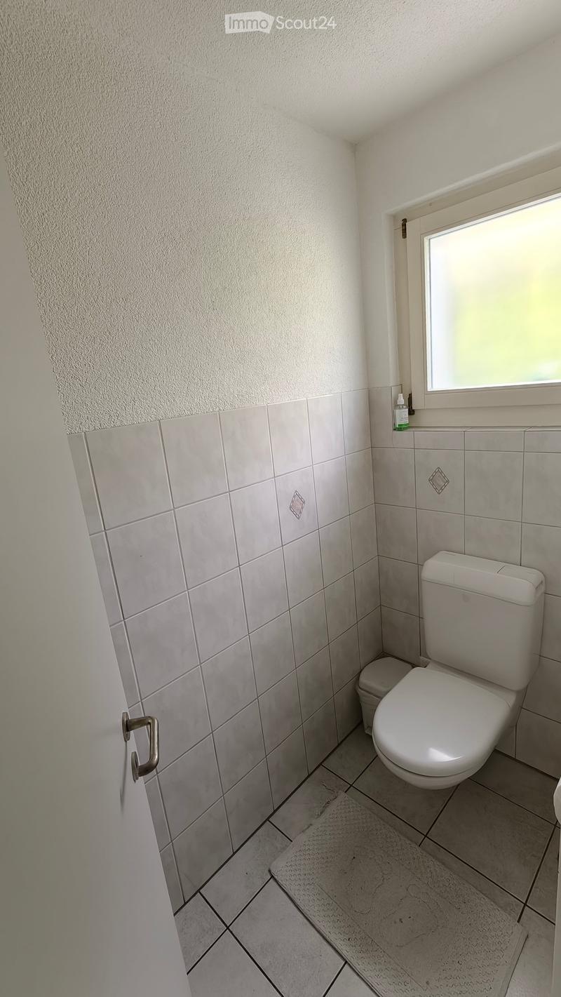 Small bathroom with white tiled walls and floor, a toilet, a small window, and a bottle on the windowsill.