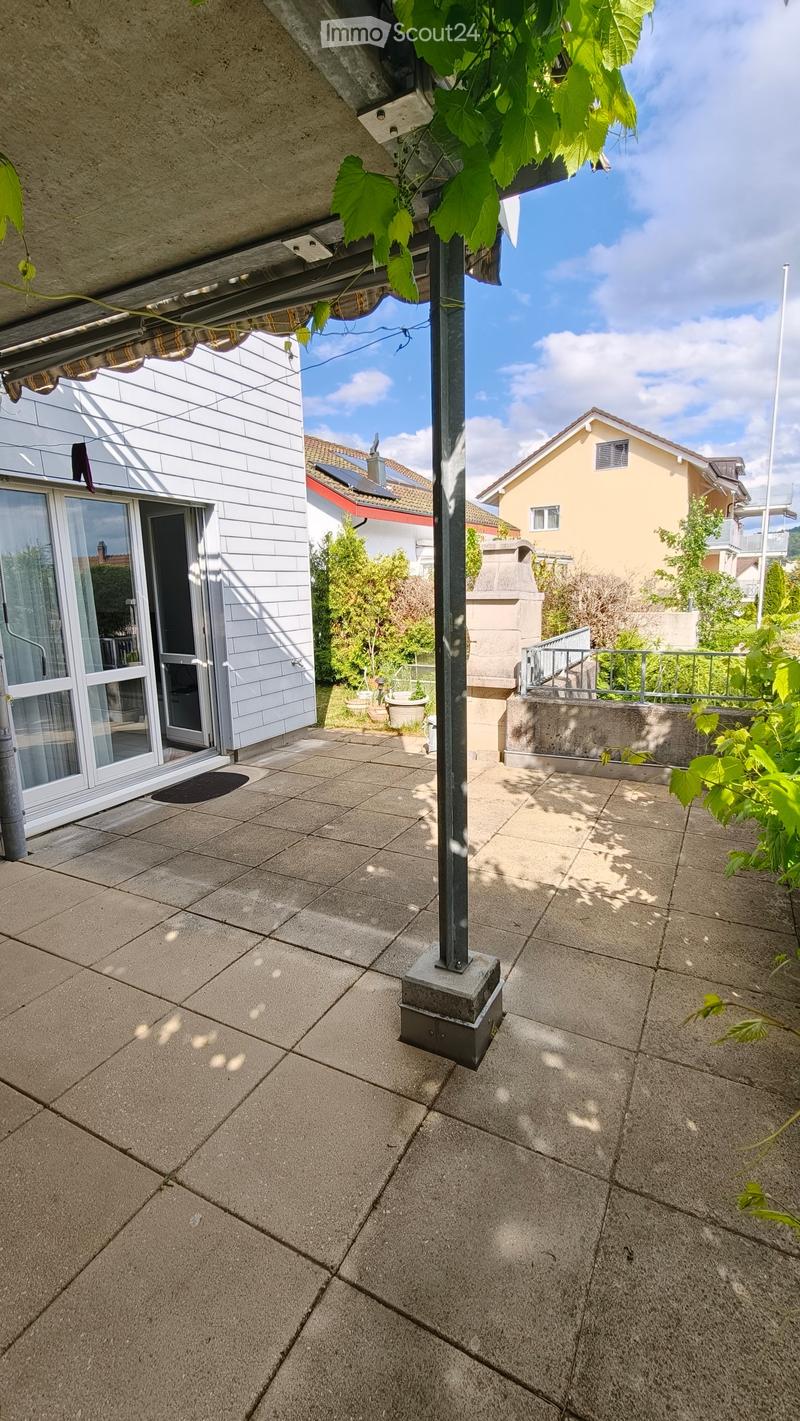 A tiled balcony area with a metal pole, glass door leading to a house, and surrounding greenery.