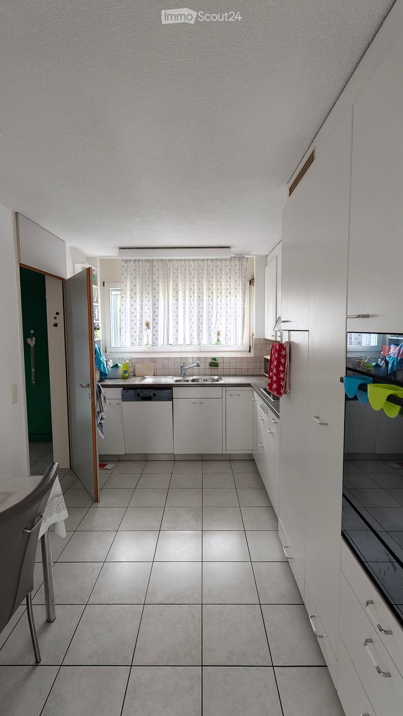 Modern kitchen with white cabinets, tiled floor, sink, dishwasher, oven, and a window with curtains. A chair is positioned near the entrance.