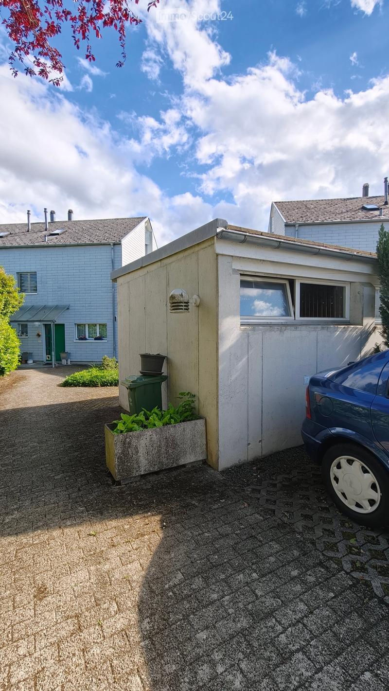 A small garden area with a concrete planter, green trash bin, and a parked car. The background shows a residential house with a grey roof and white walls.