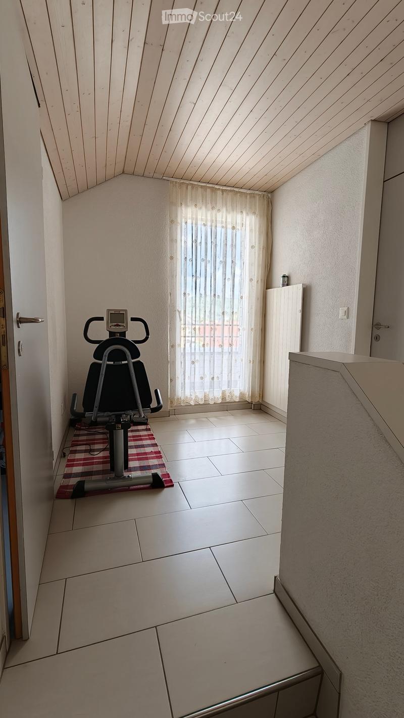 Small room with a slanted wooden ceiling, tiled floor, exercise bike, and a window with white curtains.