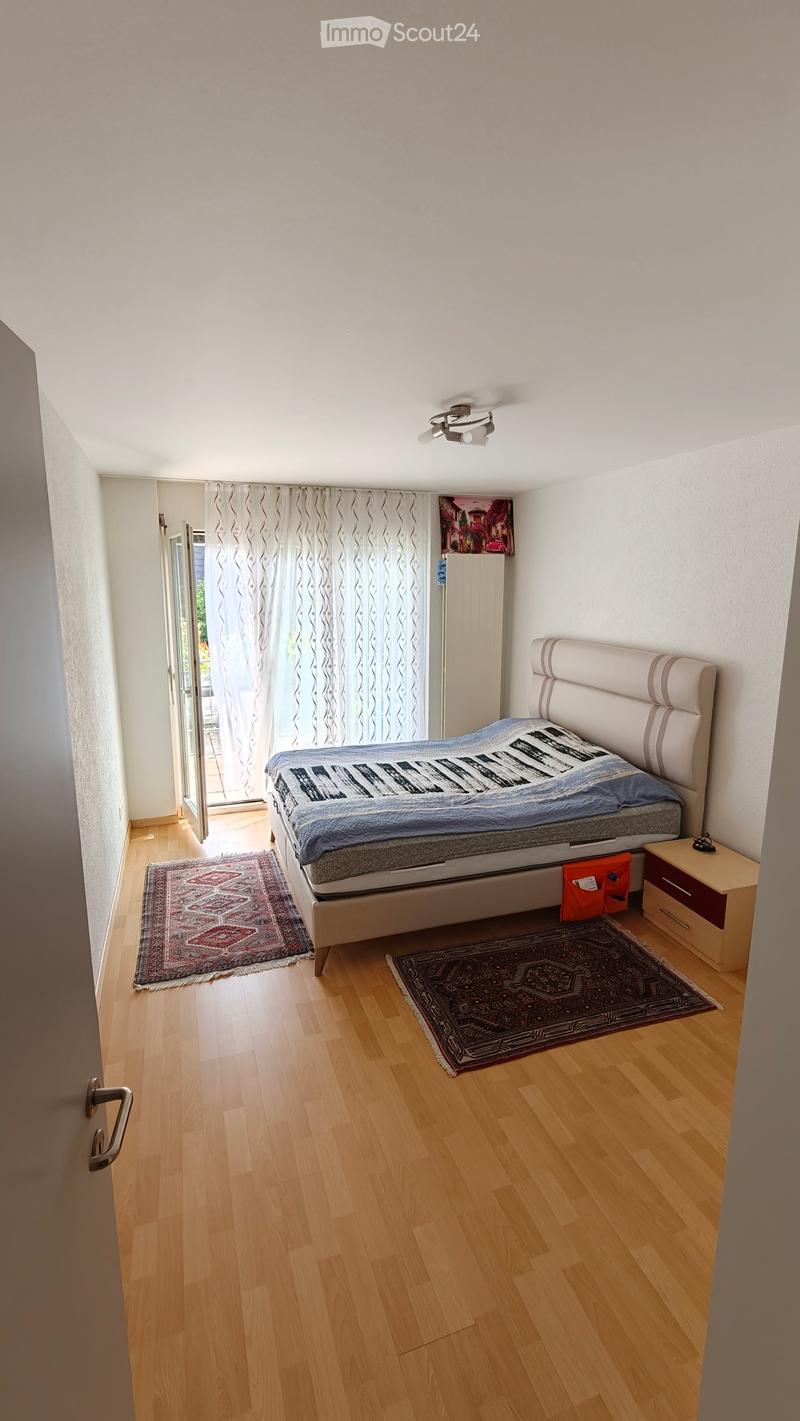 Bedroom with wooden flooring, a large bed with a headboard, two rugs, a nightstand, and a curtained window leading to a balcony.