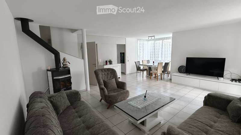 Modern living room with a grey sectional sofa, armchair, coffee table, flat-screen TV, dining area with table and chairs, and a black fireplace.