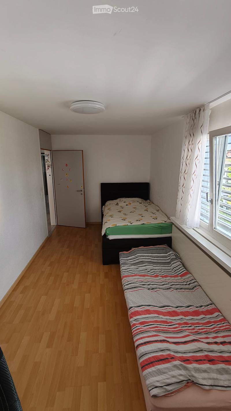 Small bedroom with wooden flooring, two beds (one full-size and one daybed), white walls, and a window with curtains.