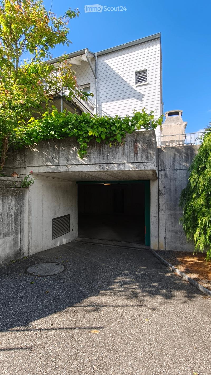 Modern two-story building with a concrete garage entrance, surrounded by greenery and a raised ground floor.