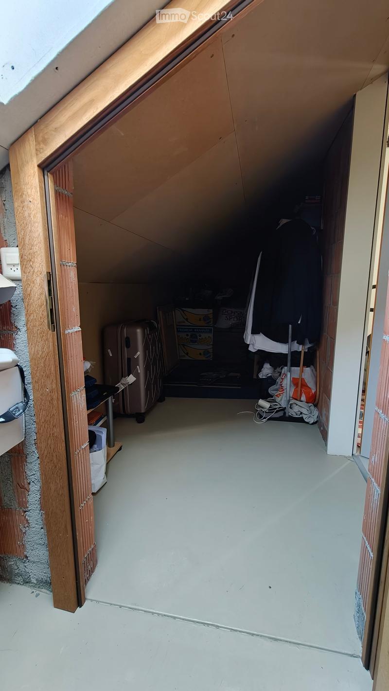 Empty attic space with slanted wooden ceiling and white walls, showing minimal furnishings and storage items.