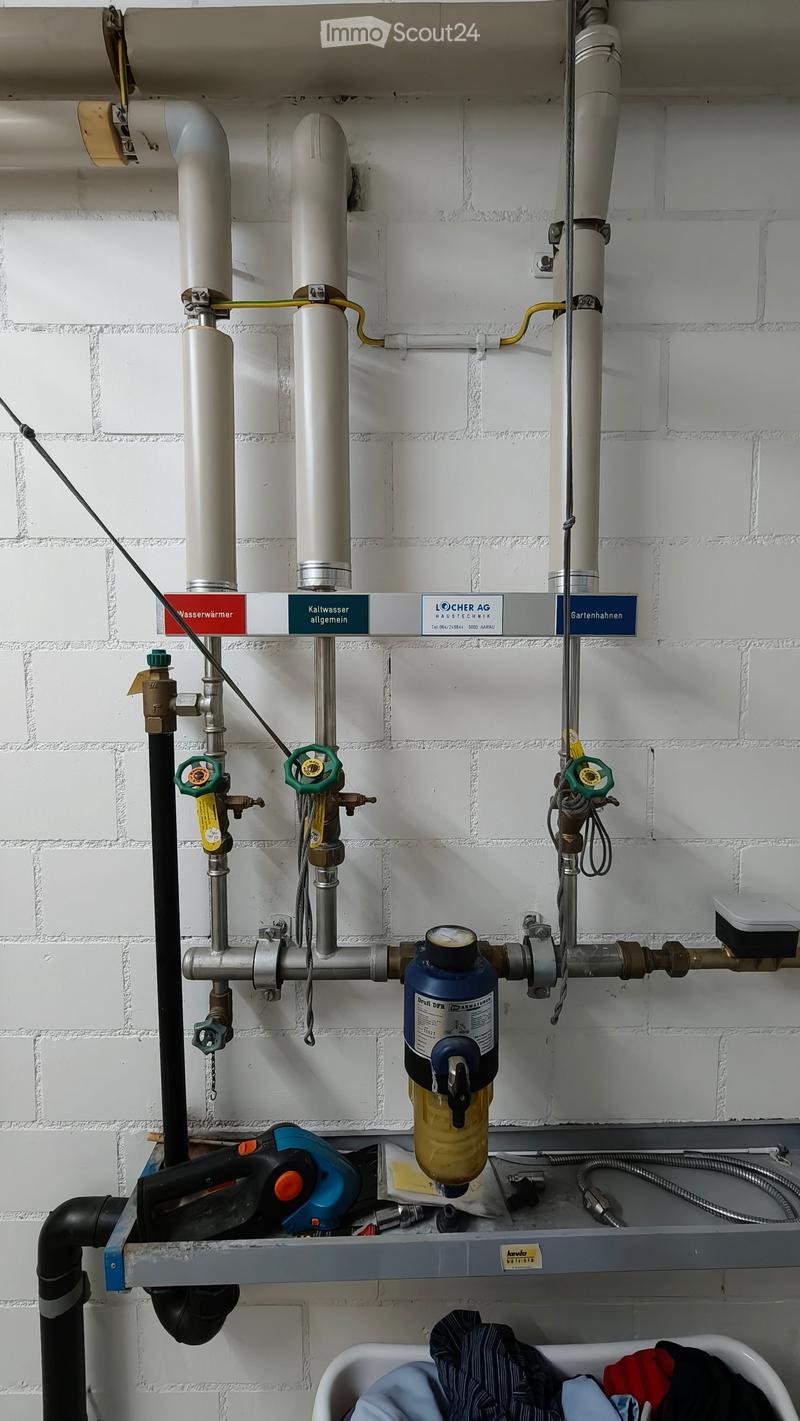 Water heating system with pipes, valves, and a filter, mounted on a white brick wall. Includes a small sink with clothes inside.