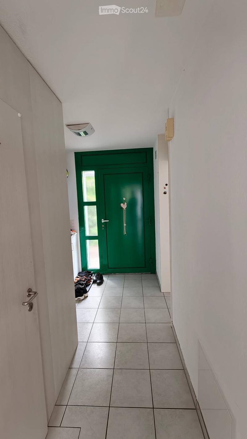 A narrow hallway with tiled flooring, white walls, and a green door at the end. A pair of shoes is placed near the door.