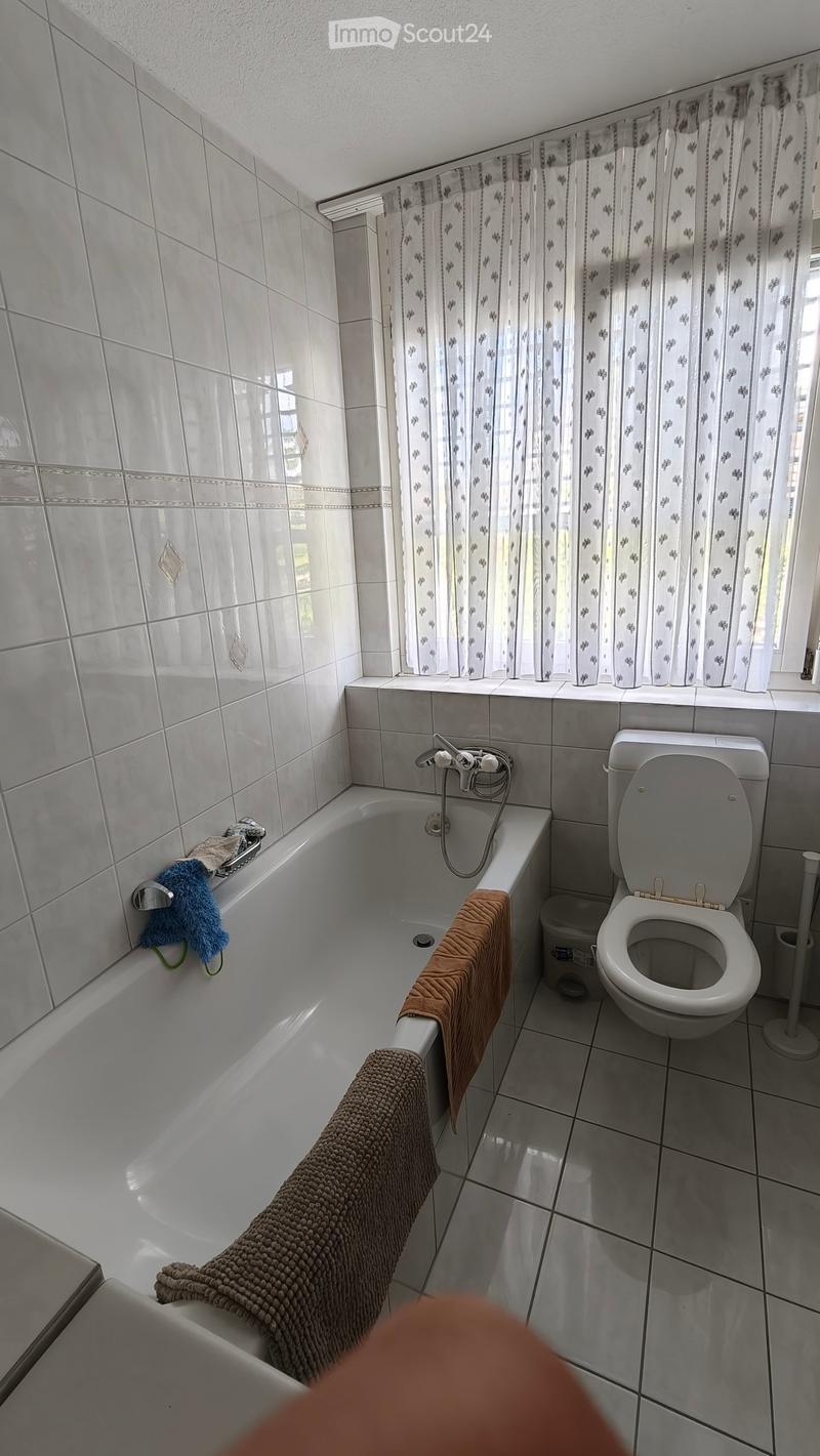 Bathroom with white tiled walls and floor, bathtub with towels, toilet, and a window with a curtain.