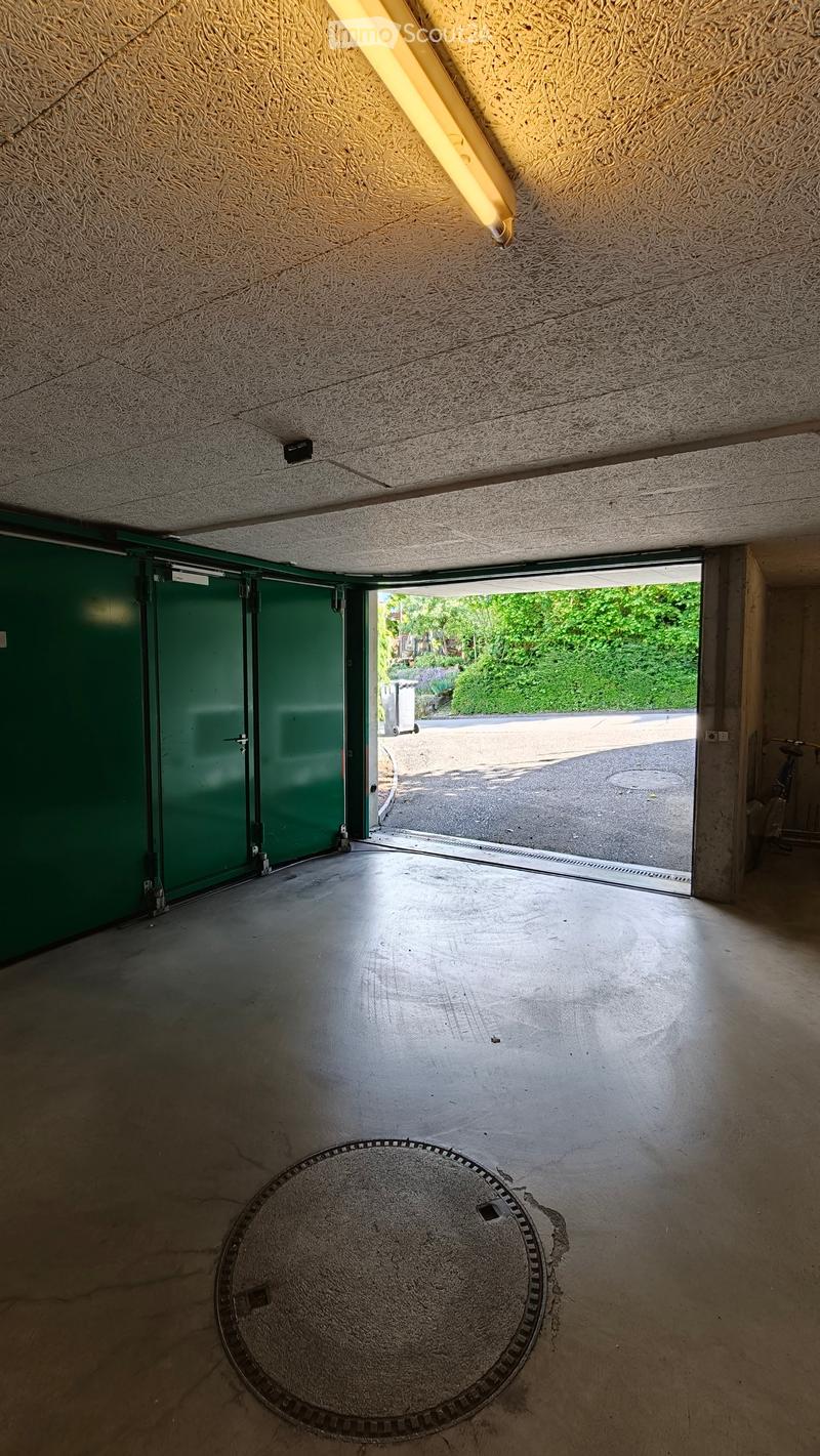 A concrete corridor with a green garage door on the left, an open doorway leading outside, and a manhole cover on the floor.