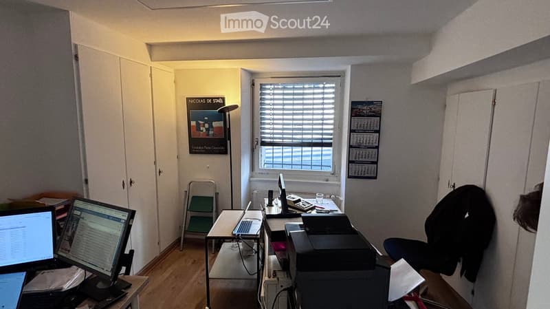 Office space with wooden flooring, two desks, laptops, printer, chair, calendar, and artwork on the wall.