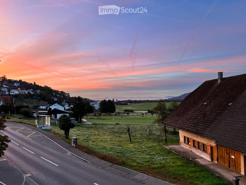 A rural house with a tiled roof and wooden shutters, situated on a grassy field near a road, with a scenic sunset view of hills and scattered houses in the background.