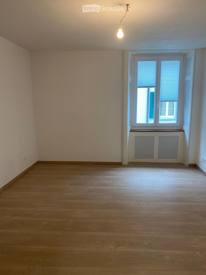 Empty room with wooden flooring, light-colored walls, and a window with blinds. A radiator is visible below the window, and a single light fixture hangs from the ceiling.